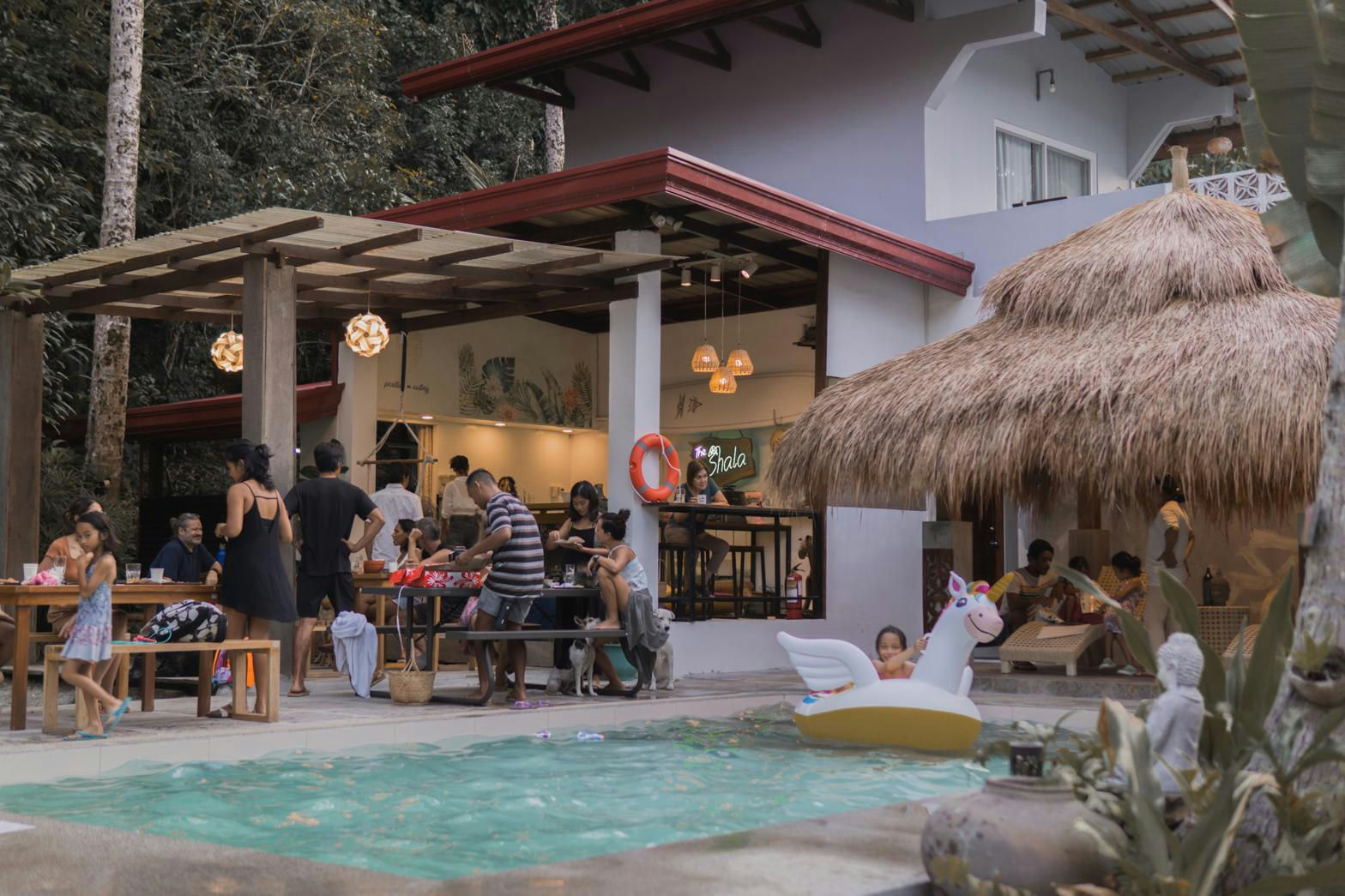 Guests gathered at wooden tables under a pergola beside the pool at Kahana Baler, with tropical greenery around