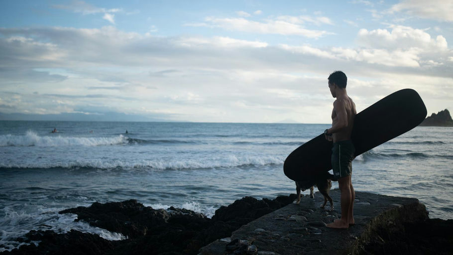 A surfer in a wetsuit holds his board on the rocky shoreline near Cobra Reef in Cemento, Baler, watching the swell before paddling out.