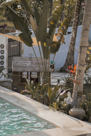 A lush tropical garden with a wooden hut, banana trees, and the edge of a swimming pool at Kahana Baler in Cemento.